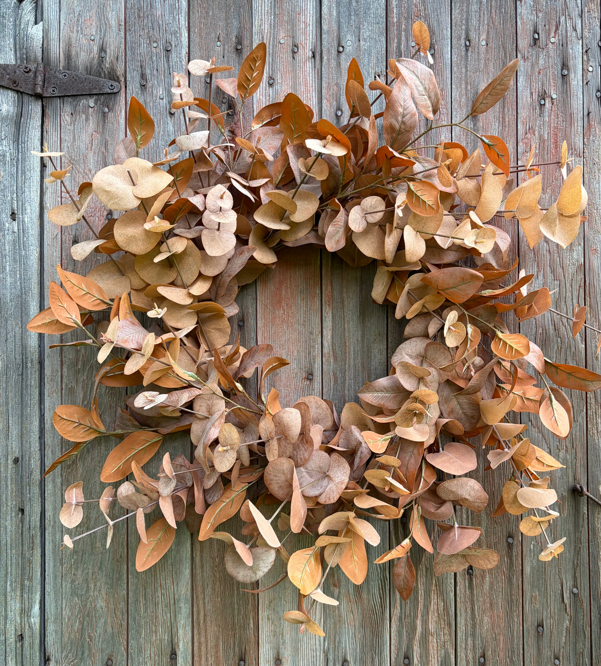 A rustic dried eucalyptus wreath in warm brown, tan, and beige tones, displayed against a weathered gray wooden door.