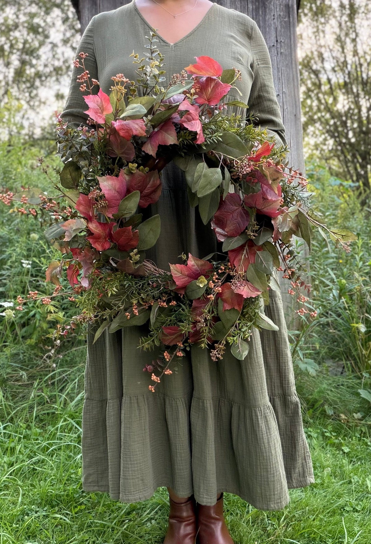 A person wearing an olive green long-sleeve V-neck dress stands outdoors in front of a wooden post, holding a large autumn wreath with red and burgundy leaves, green foliage, and small orange berries.