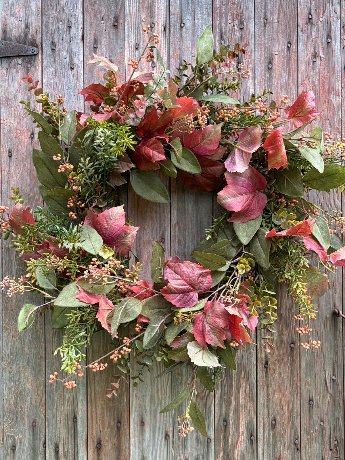 An autumn-themed floral wreath with red and pink leaves, green foliage, and small pink berries, displayed against a weathered gray wooden door.