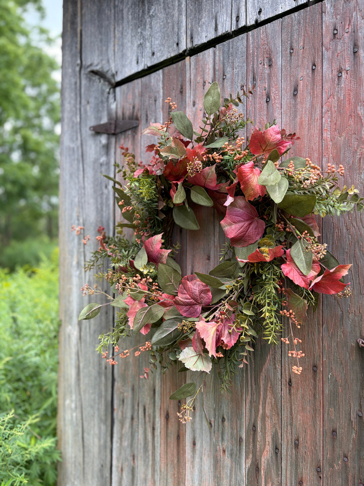 An autumn leaf wreath with red, burgundy, and green foliage hanging on a weathered brown wooden barn door.