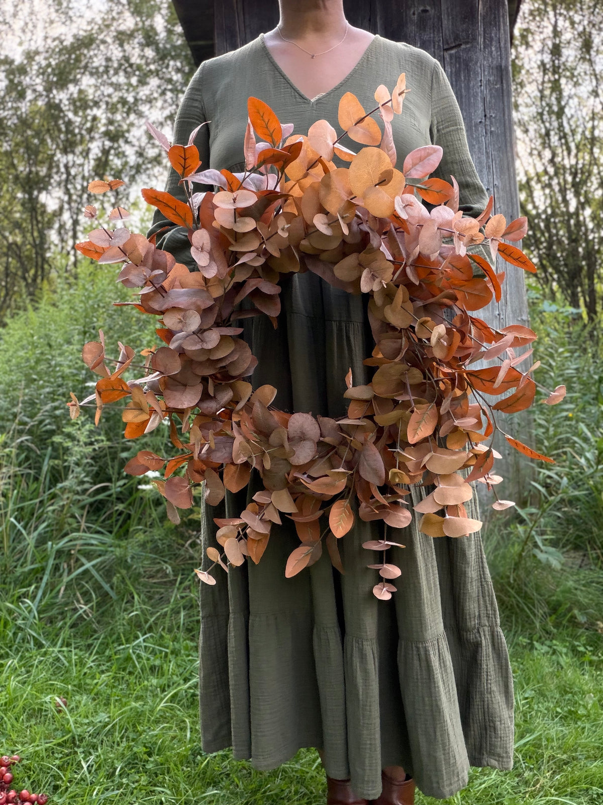A person wearing an olive green dress holds a large eucalyptus wreath with dried autumn leaves in shades of brown, orange, and beige, set against a grassy outdoor background.