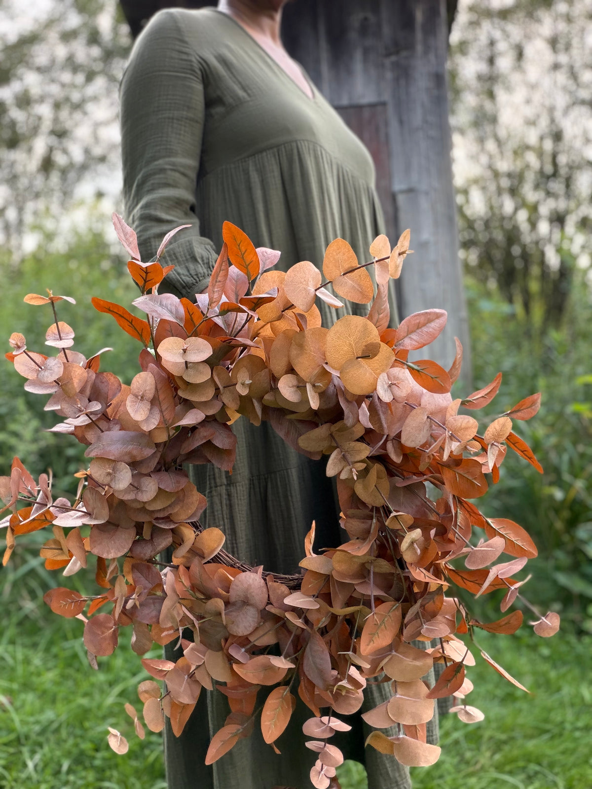 A person outdoors holding a large decorative wreath made of dried eucalyptus branches in muted orange, tan, and dusty rose tones.