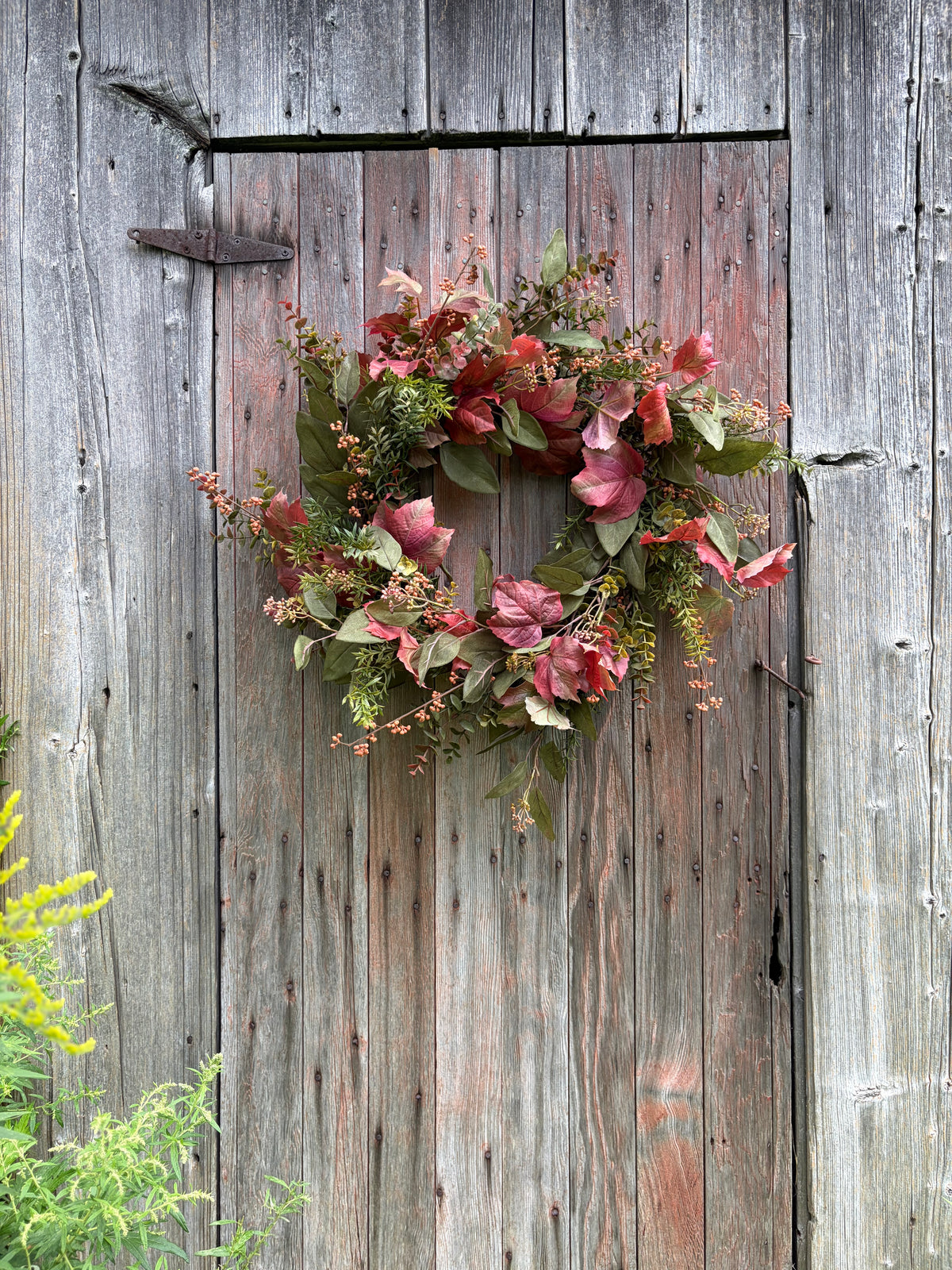 An autumn-themed floral wreath with pinkish-red leaves and green foliage, hanging on a weathered gray wooden barn door.