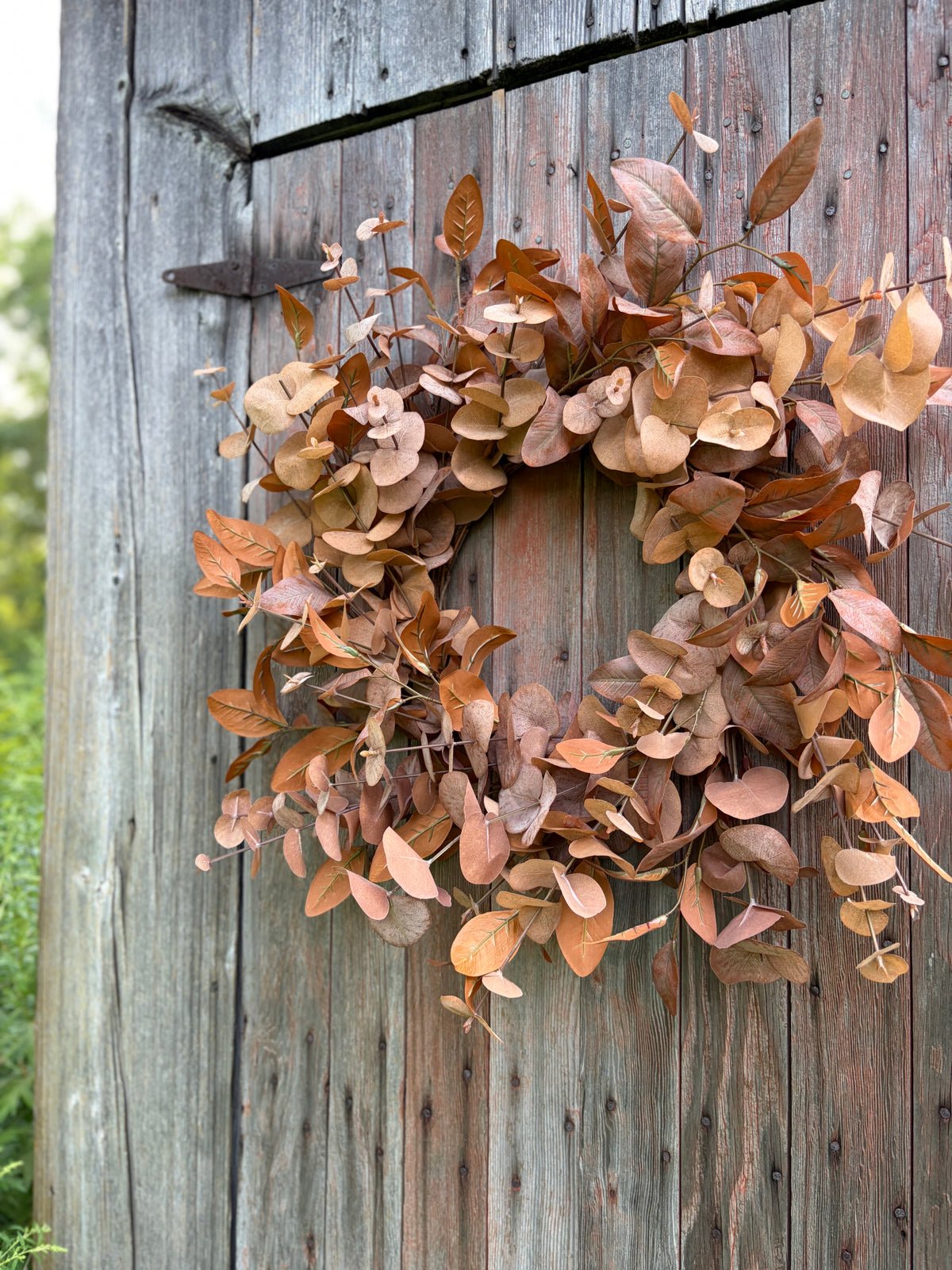An autumn eucalyptus wreath with dried brown, tan, and muted orange leaves, hanging on a weathered gray wooden door.