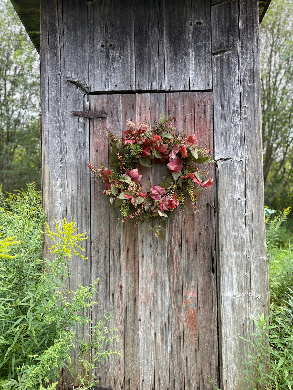 A rustic artificial floral wreath with pink and red flowers and green foliage hanging on the weathered wooden door of a small shed, surrounded by green garden plants.